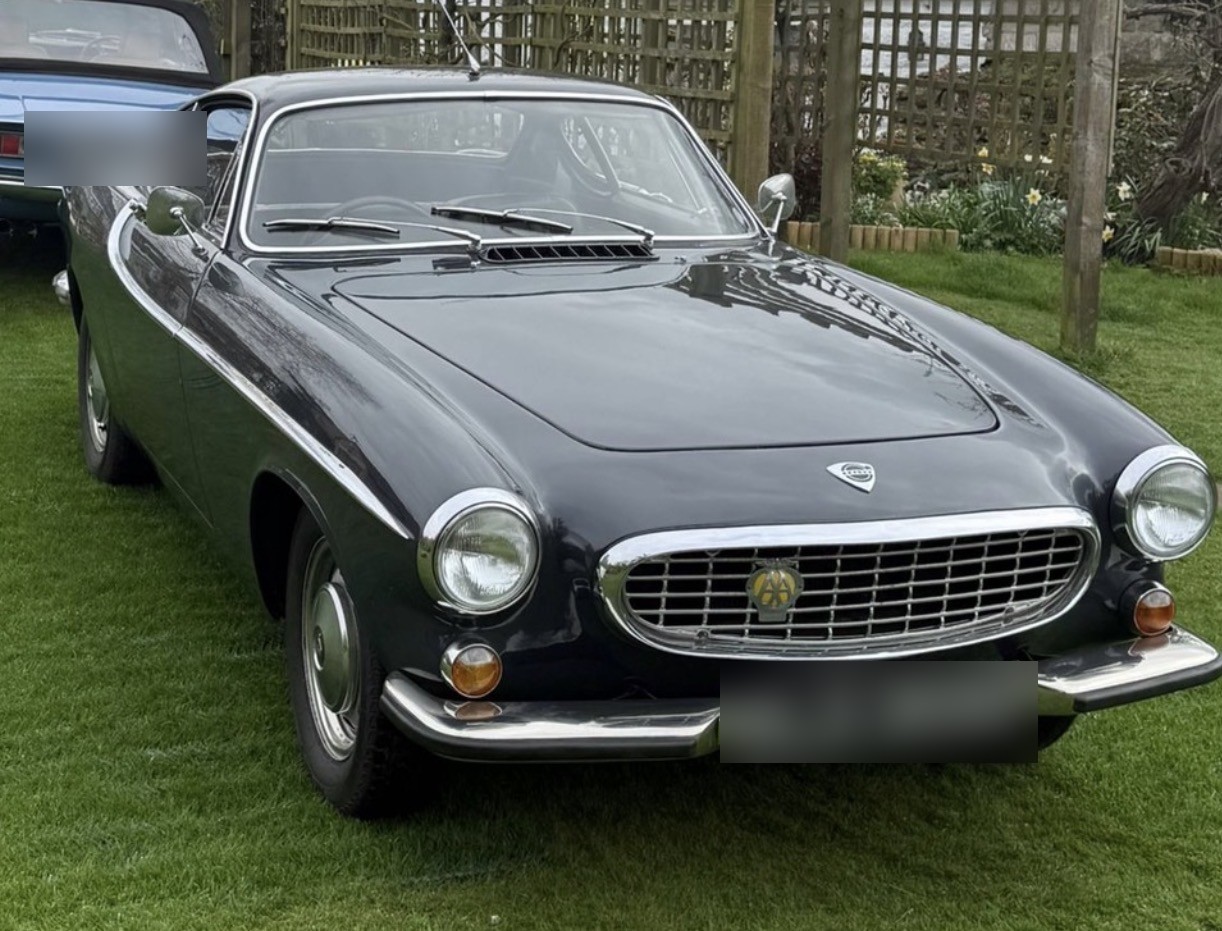 A dark grey Volvo P1800S coupé on a lawn, an AA badge in the grille