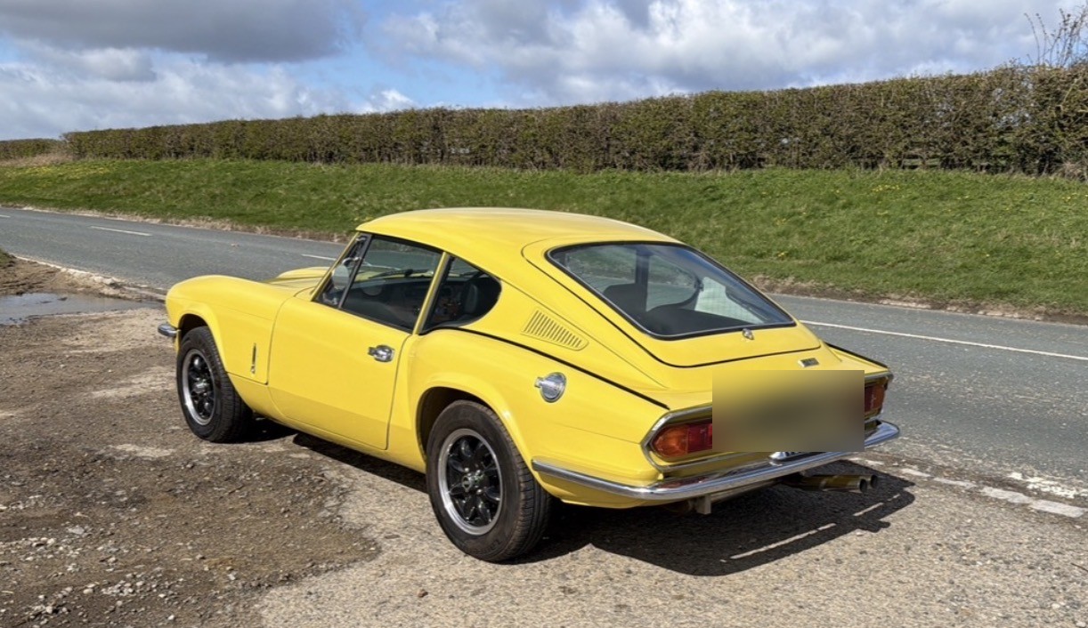 A yellow Triumph GT6 Mk3 seen from the rear three-quarter, parked on the verge of a country lane beside a hedge