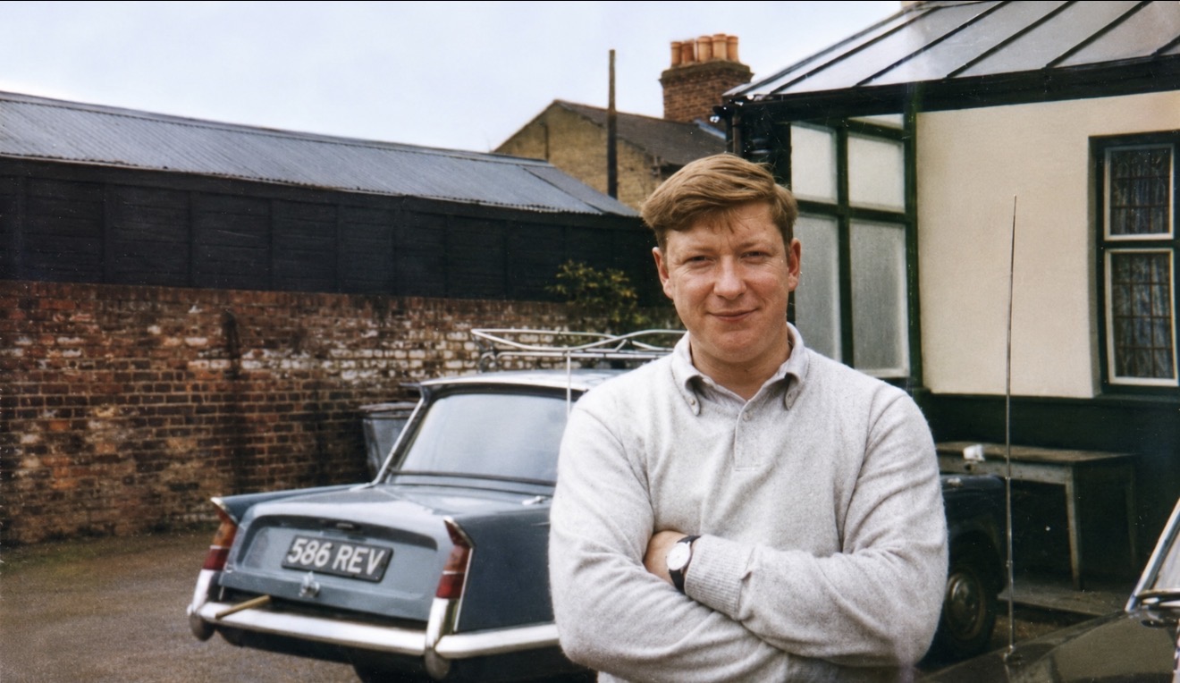 A young man in a pale jumper, arms folded, standing on a yard in front of a Triumph saloon registered 586 REV, with a brick wall and corrugated shed behind him