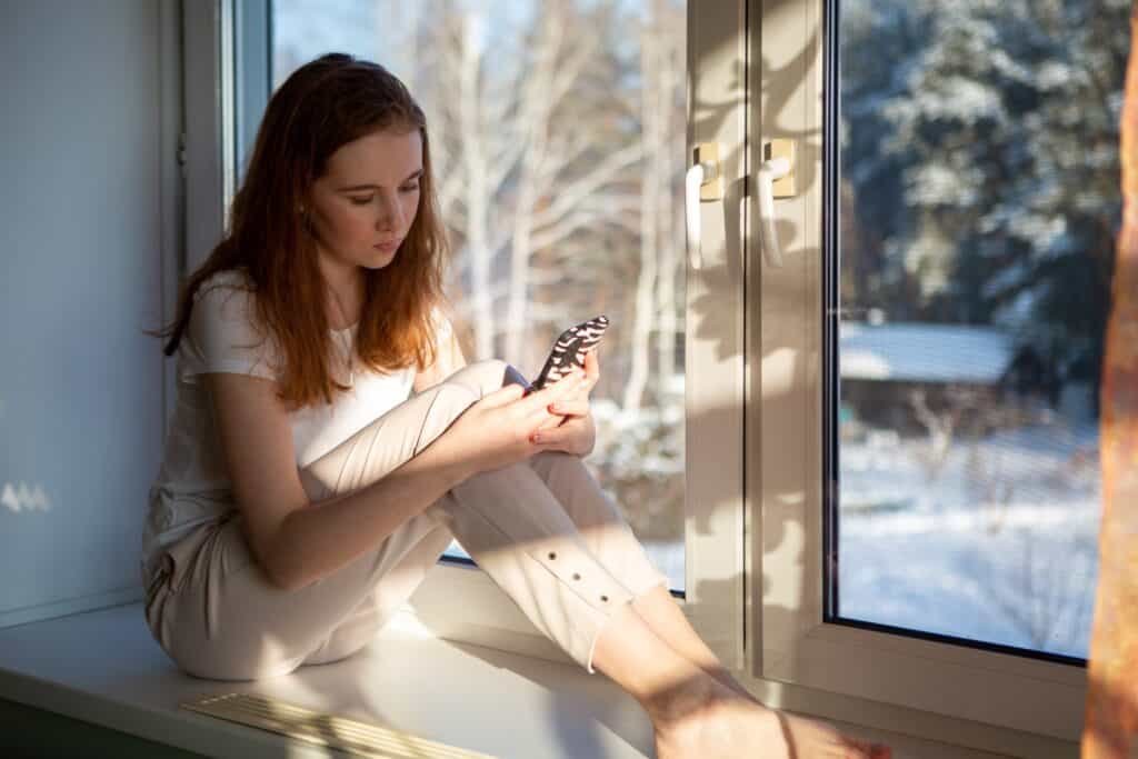 A girl on a window ledge looking at her smart phone in deep thought