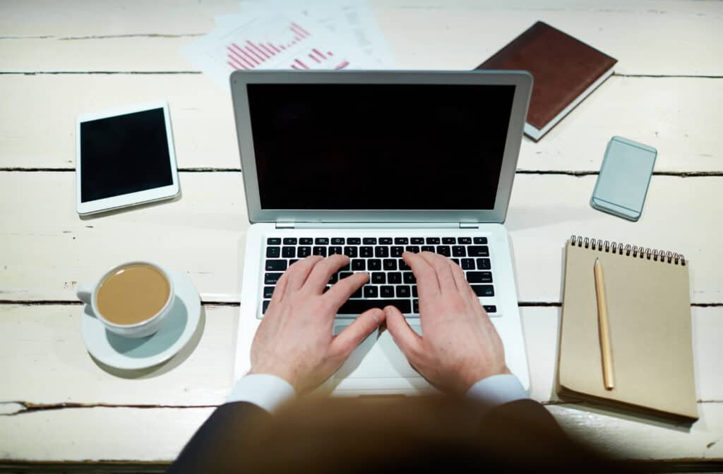 A man wirkig on a wooded desk with his laptop, along with a tablet, note pad, smart phone and coffee