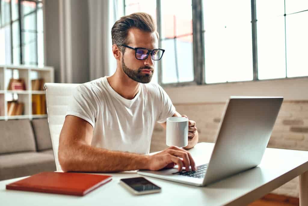 Man in glasses with a mug of coffee works at a laptop checking web addresses