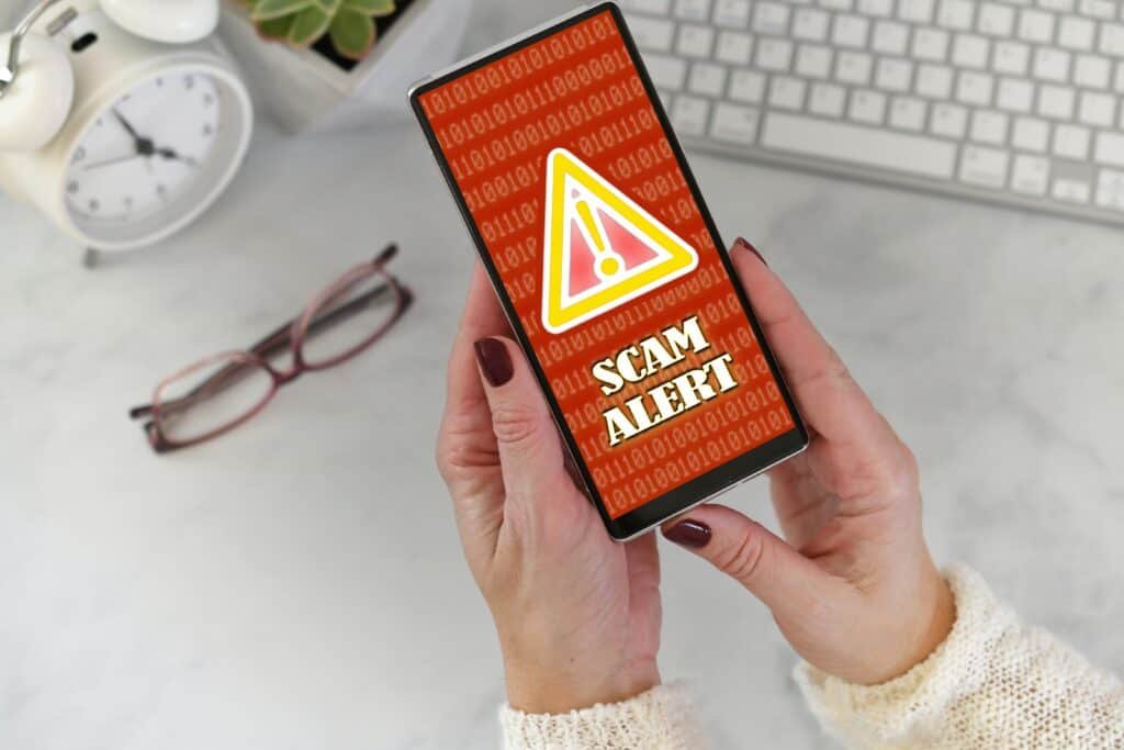 Woman holding a phone with a red screen and a yellow triangle with an explanation mark in the middle stating Scam Alert with a red background