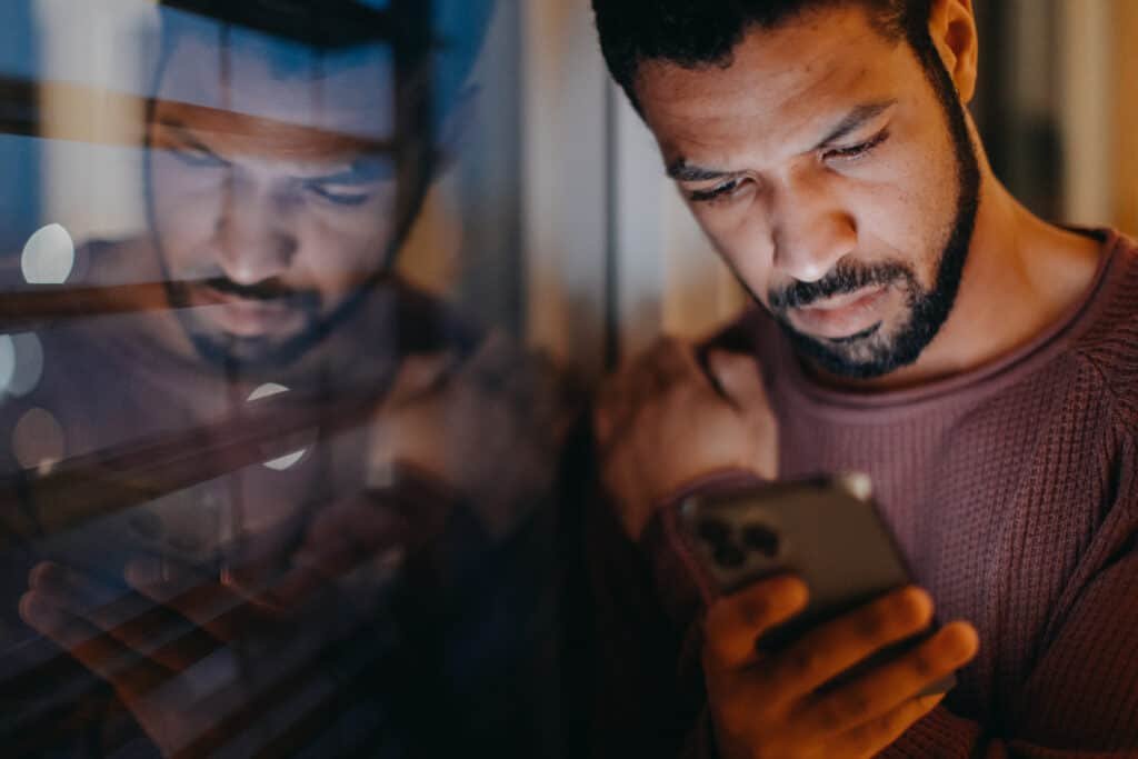 A young man with a smart phone looks intense, standing by window in evening at home.