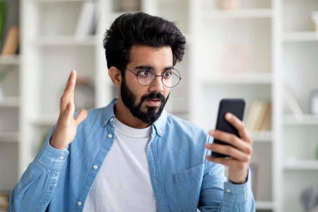 Bad News. Shocked Man Looking At Smartphone In His Hand, Furious as he has been Scammed Online
