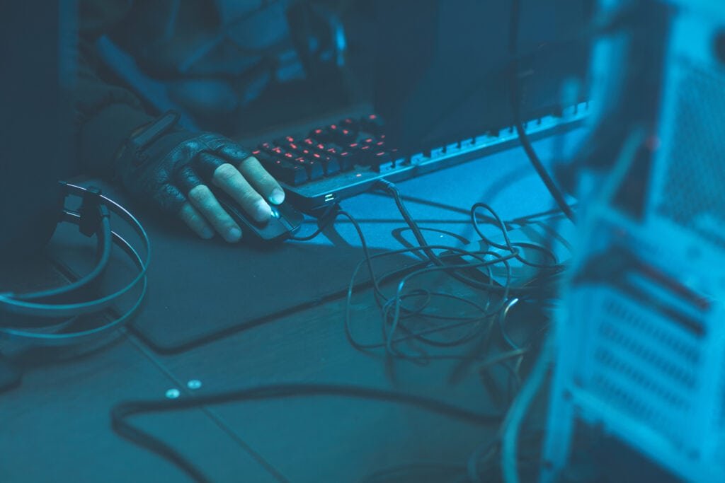 What is vishing - Close-up of unrecognisable male cybercriminal in fingerless gloves sitting at desk with wires and cables and using mouse while hacking computer system