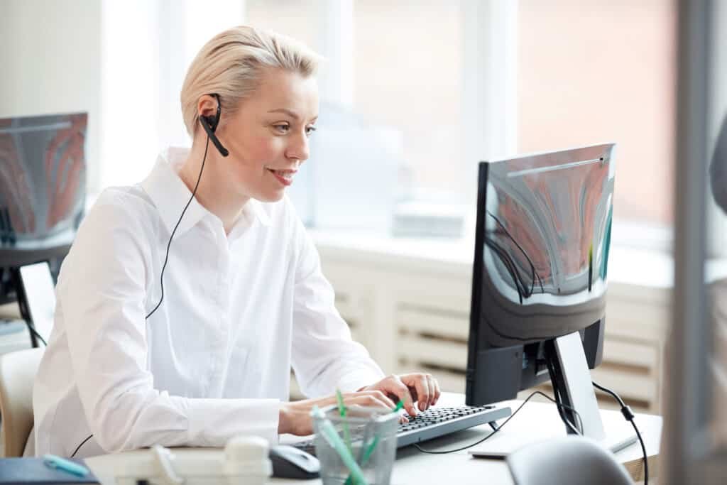 Side view portrait of female online banking operator using laptop and wearing headset while performing customer support service