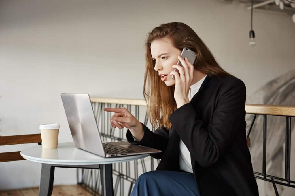 Girl picking item in online-store. Indoor shot of good-looking female student in cafe, sitting over laptop, pointing at screen while talking on smartphone, wanting to buy something in internet.