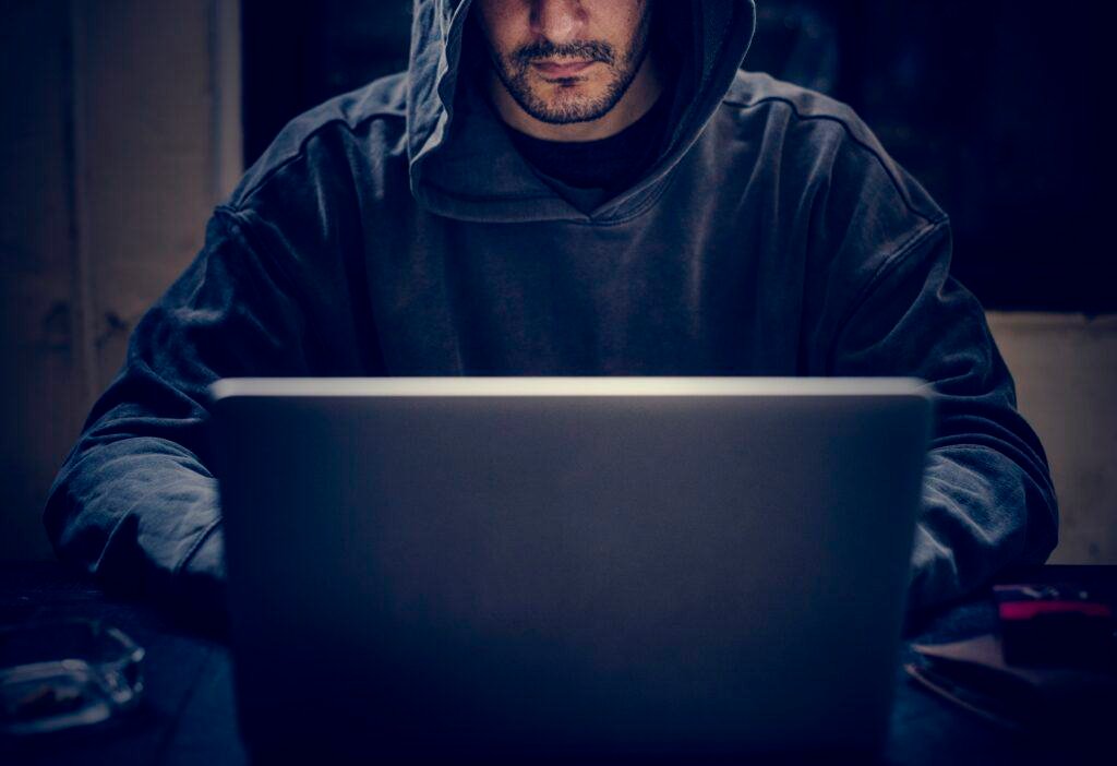 A hooded cybercriminal on a laptop scaming victims using phishing scams