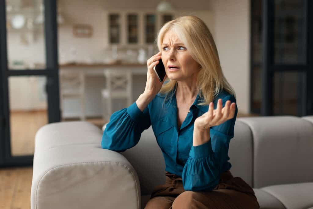 Frustrated mature woman talking on smart phone at home, sitting on sofa.