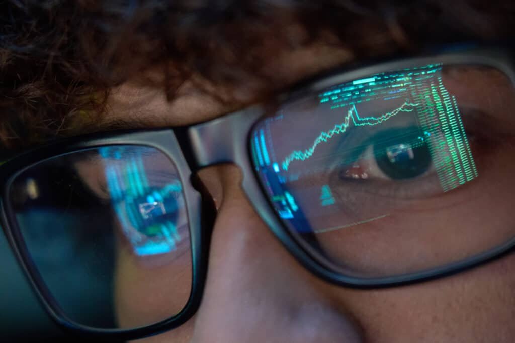 Young man wearing glasses looking at computer screen with trading charts reflecting in eyeglasses watching stock trading market financial data growth.