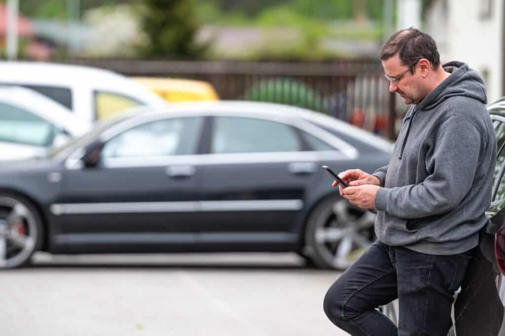A man with smart phone standing next to the car, using his phone to check for parking app scams.