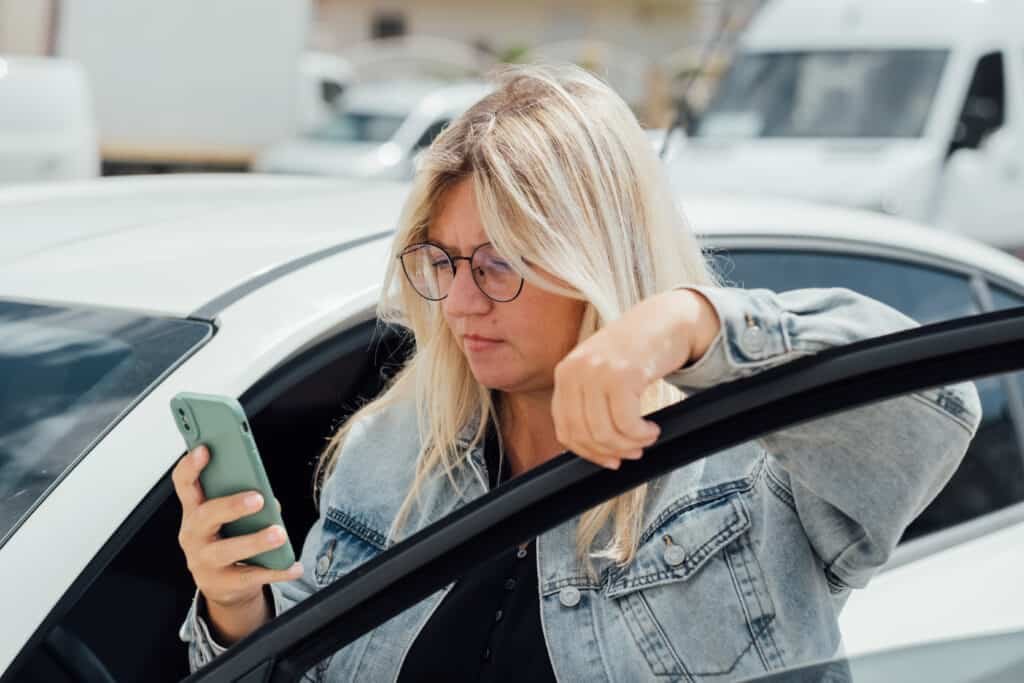 Woman standing near car and looking at her mobile phone, paying for parking and navigates the city, checking to see if she has become a victime of a Parking App Scam.