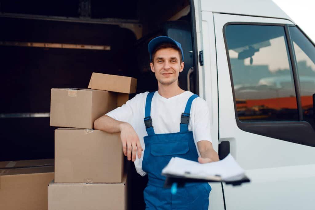 Deliveryman in uniform holding parcel and notebook, carton boxes by a van, delivery service. Man standing at cardbord. This is part of a parcel delivery scamming process