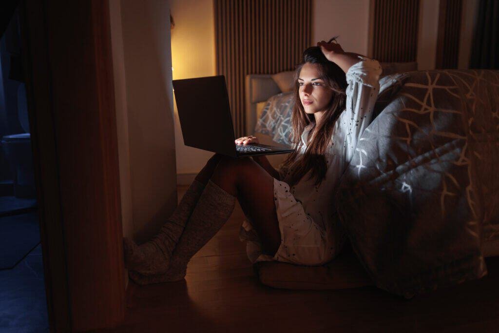 Beautiful young woman sitting on the floor by the bed, holding laptop computer in her lap, chatting using online dating site