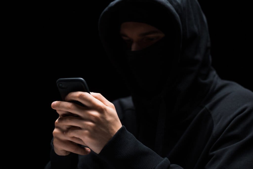 selective focus of hacker in mask using smartphone isolated on black