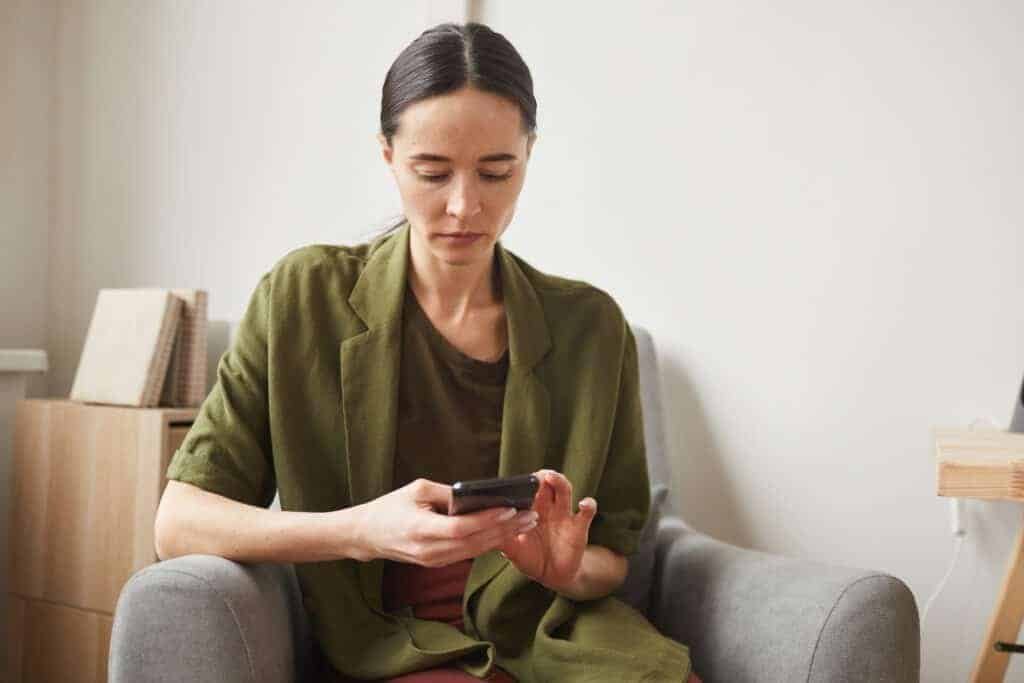 An adult woman using smartphone while relaxing in armchair, realising her facebook accont hacked phone and email changed