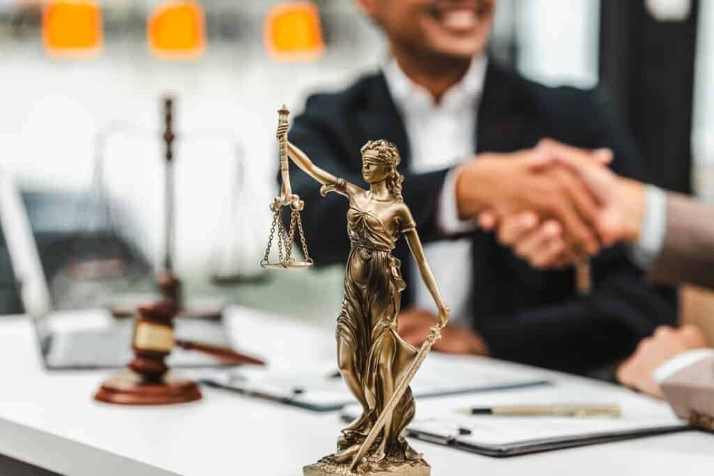 A lawyer shaking hands with thier client over a white desk with a legal image justice.